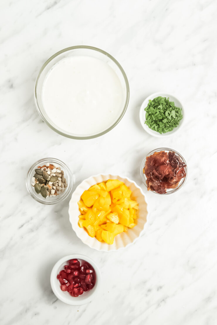 Overhead view of bowls f ingredients needed to make coconut yogurt parfait including coconut yogurt, mixed berries, frozen mango, dates, walnuts, pumpkin seeds, sunflower seeds, pomegranate arils, and chopped mint.