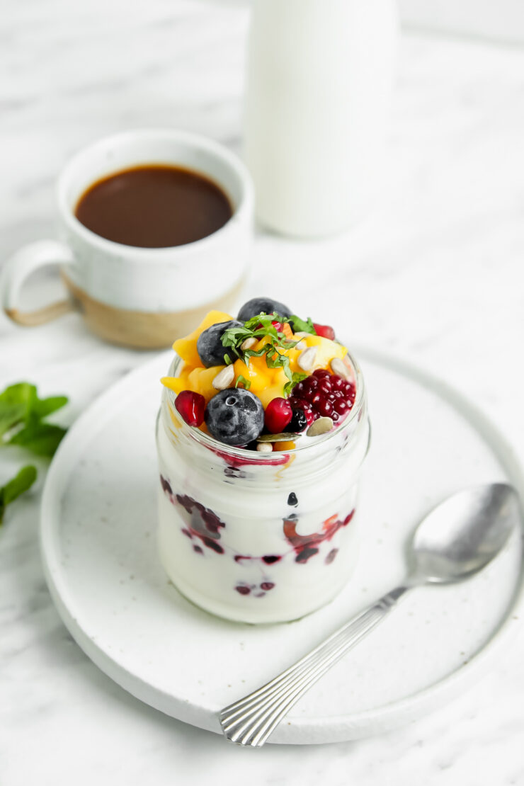 Overhead shot of a jar of coconut yogurt parfait, which is sitting on top of a white plate and has a spoon on the side. It includes layers of fresh fruit and coconut yogurt along with a mixture of fresh berries, thawed frozen mango, and sliced mint on top.