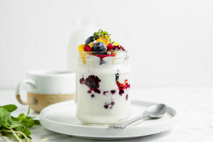 Jar of coconut yogurt parfait sitting on a white plate with a white and brown mug sitting behind and mint sitting off to the side. The parfait includes layers of coconut yogurt and berries with a pile of fresh berries, mango, and mint on top.