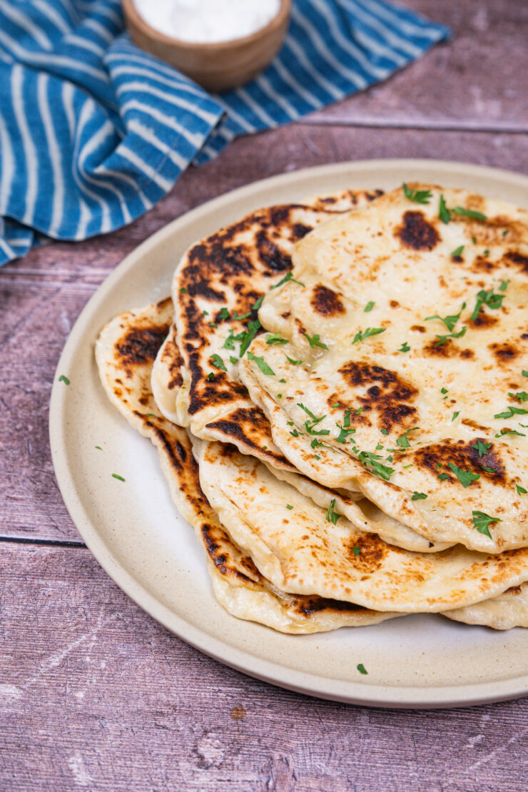 Stack of vegan naan on a white plate with chopped cilantro sprinkled on top. A blue and white kitchen towel with a small brown bowl on top is sitting behind.