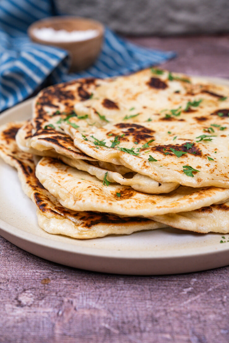 Closeup view of a stack of vegan naan on a white plate and sprinkled with chopped cilantro. A small brown bowl and white and blue kitchen towel is sitting behind it.