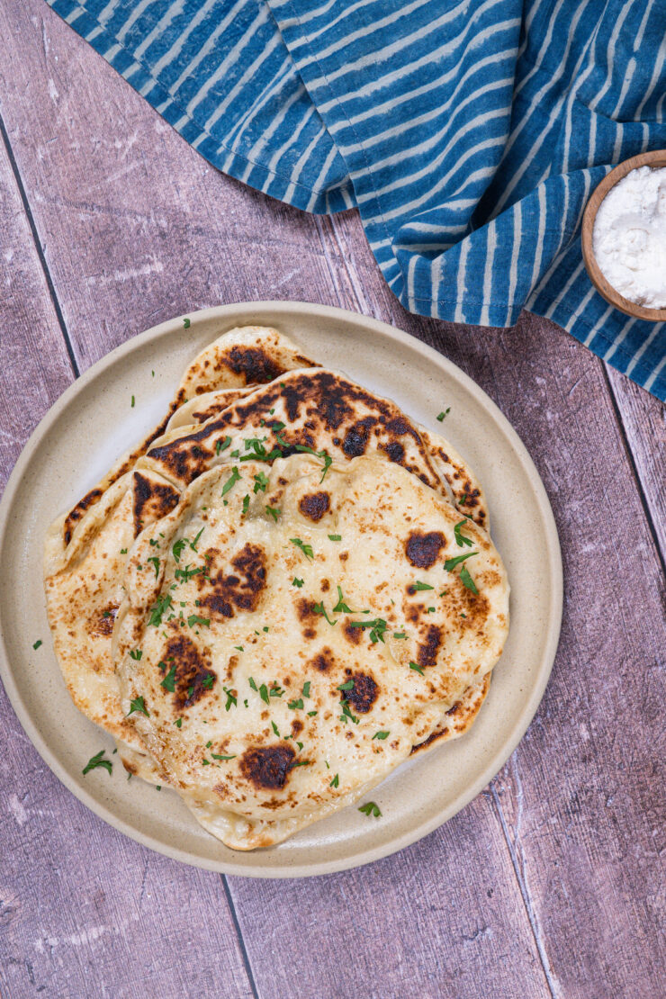 Closeup view of a stack of golden vegan naan on a white plate with cilantro sprinkled on top. A blue and white striped kitchen towel is sitting in the top right corner with a small wooden bowl of flour on top.