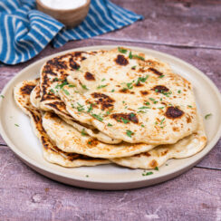 Stack of vegan naan bread on a white plate with chopped cilantro sprinkled on top. A blue and white striped towel and a small brown bowl are sitting behind it.