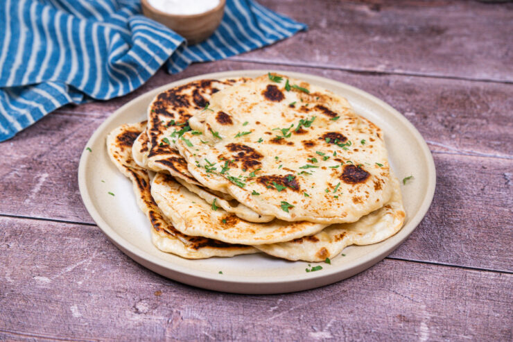 Stack of vegan naan bread on a white plate with chopped cilantro sprinkled on top. A blue and white striped towel and a small brown bowl are sitting behind it.