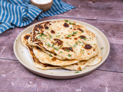 Stack of vegan naan bread on a white plate with chopped cilantro sprinkled on top. A blue and white striped towel and a small brown bowl are sitting behind it.