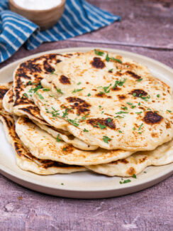 Stack of vegan naan bread on a white plate with chopped cilantro sprinkled on top. A blue and white striped towel and a small brown bowl are sitting behind it.