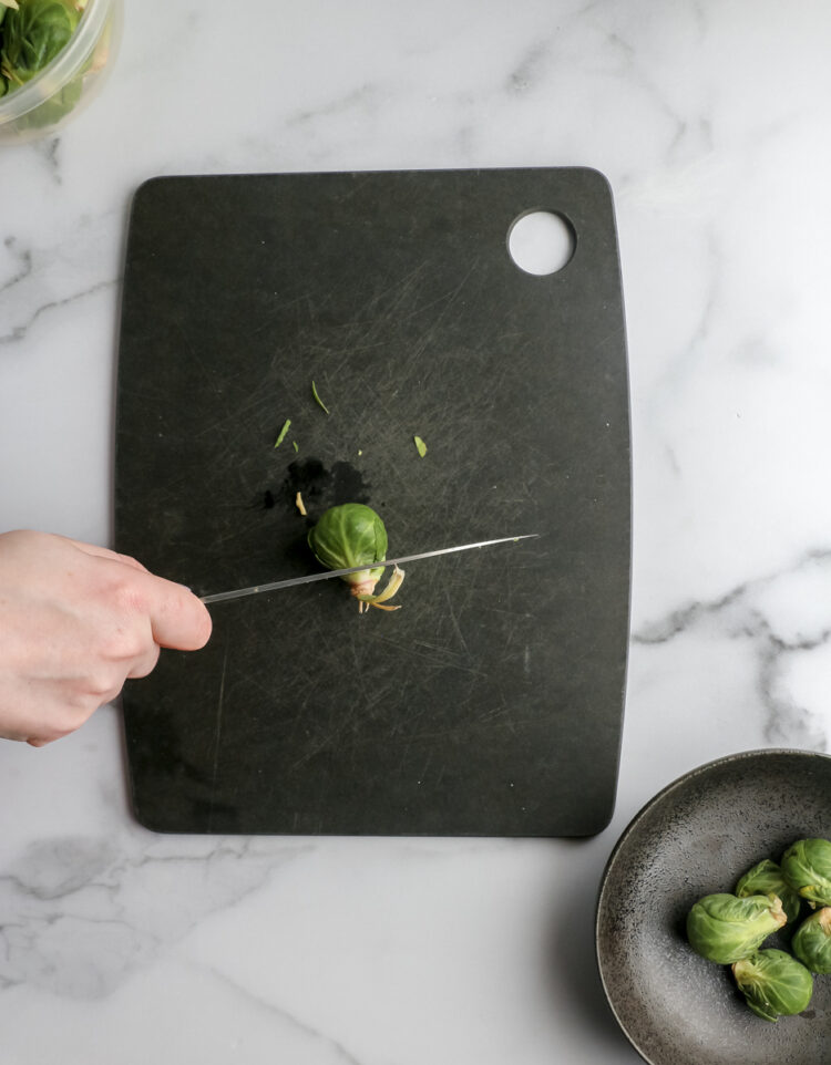 A knife slices through the stem of a brussels sprout on a chopping board.