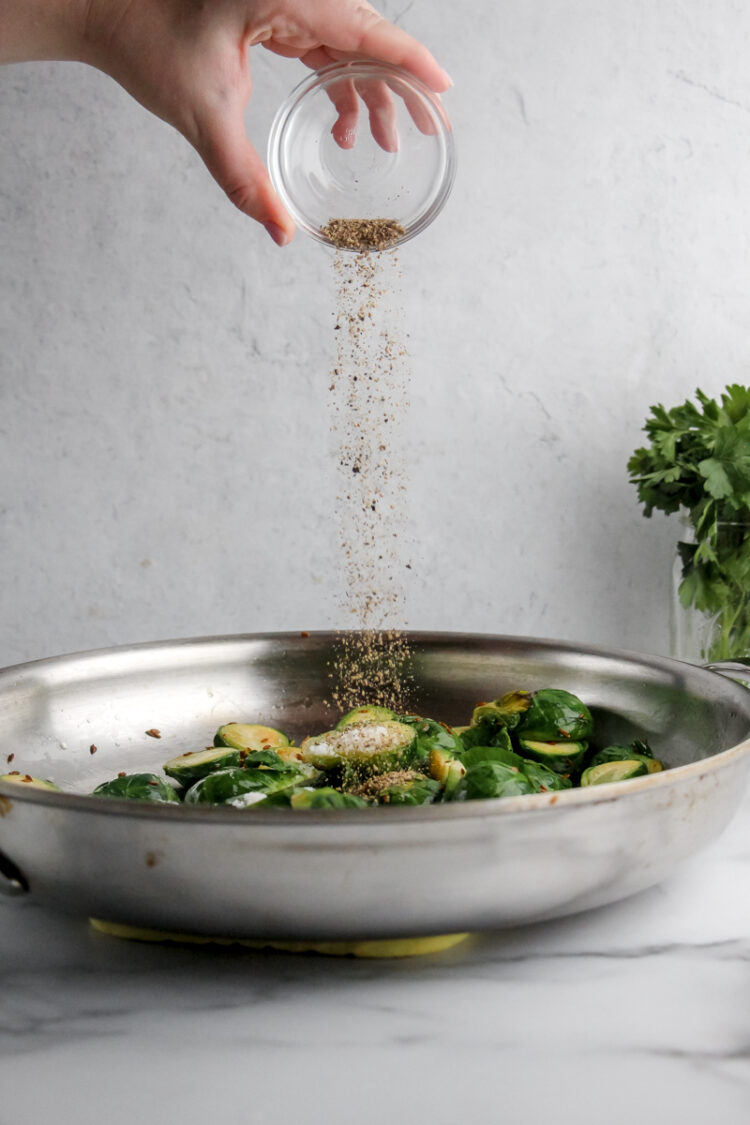 A hand pours cumin powder over a metal dish containing chopped brussels sprouts with olive oil and mustard seeds.