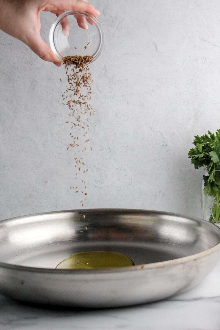 A hand pours cumin seeds into a metal dish containing olive oil.