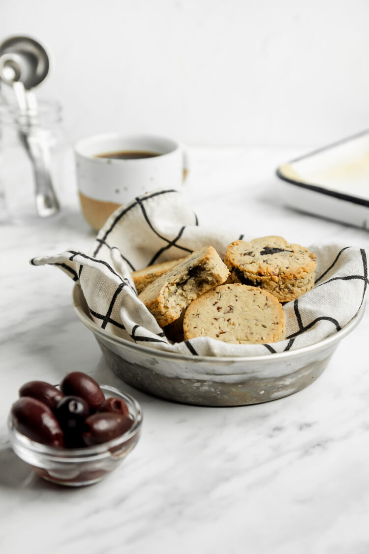 Kitchen towel-lined bowl of grain-free almond flour biscuits with a cup of coffee behind and a small bowl of black olives on the side.