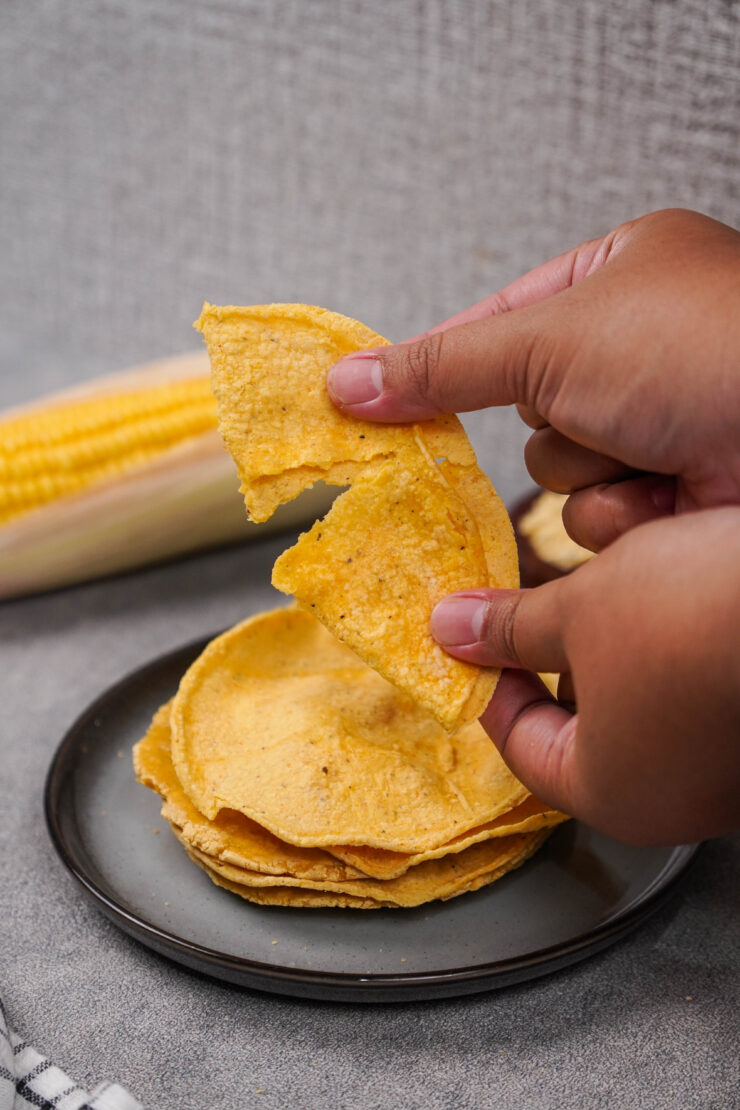 Hands break apart a masa harina tortilla, with more tortillas on a plate in the background.