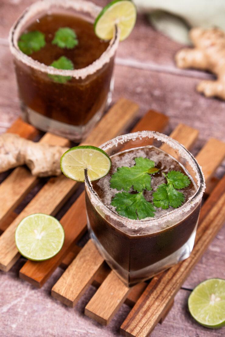 Overhead shot of two salt-rimmed square-shaped glasses filled with brown colored ginger tamarind margarita with lime slices on the rims of the glasses and whole cilantro leaves floating on top of the cocktail mixture. Lime halves and a whole ginger root are sitting on the side.