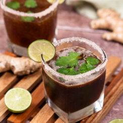 Two salt-rimmed glasses filled with brown colored ginger tamarind margarita with lime slices on the rims of the glasses and whole cilantro leaves floating on top of the cocktail mixture. Lime halves and a whole ginger root are sitting on the side.