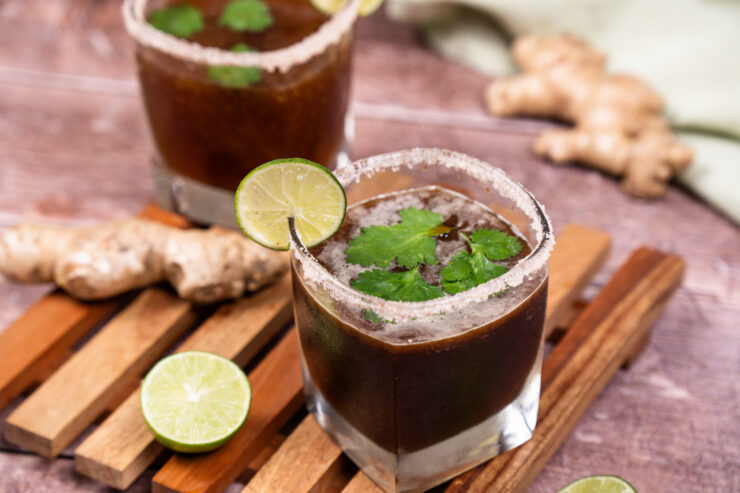 Two salt-rimmed glasses filled with brown colored ginger tamarind margarita with lime slices on the rims of the glasses and whole cilantro leaves floating on top of the cocktail mixture. Lime halves and a whole ginger root are sitting on the side.