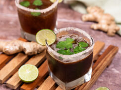 Two salt-rimmed glasses filled with brown colored ginger tamarind margarita with lime slices on the rims of the glasses and whole cilantro leaves floating on top of the cocktail mixture. Lime halves and a whole ginger root are sitting on the side.