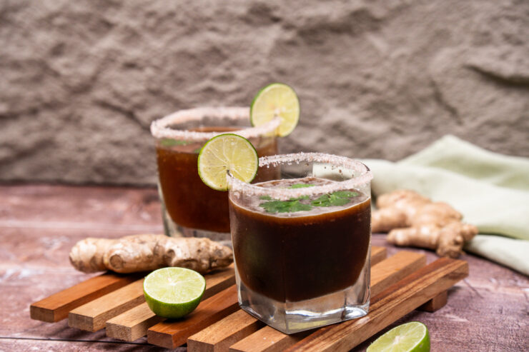 A side view of two square-shaped tumblers filled with brown colored ginger tamarind mocktail. The glasses are garnished with a lime slice and salt rim. There are lime halves and a whole ginger root off to the side.