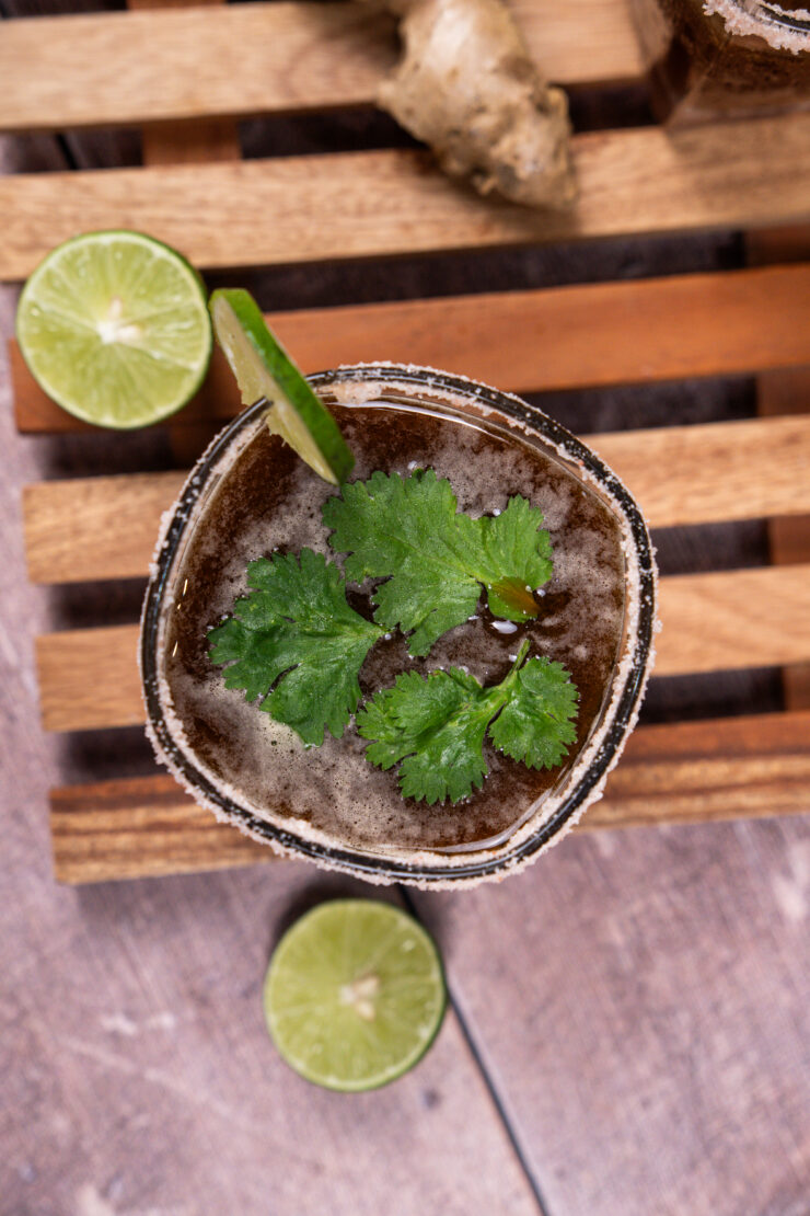 Overhead view of a tumbler glass with a salt rim that is filled with dark brown ginger tamarind margarita and has cilantro leaves floating on top. The glass has a lime slice on the rim.