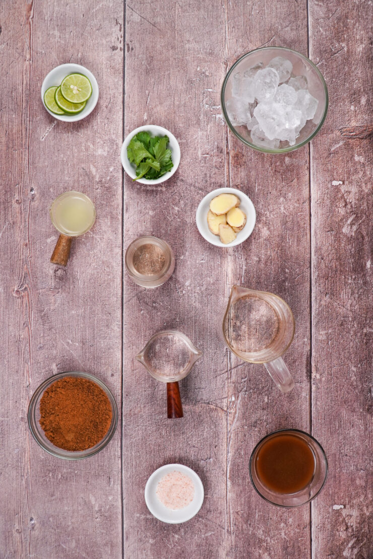 Overhead shot of bowls of ingredients needed to make this ginger tamarind margarita. It includes small bowls of lime slices, cilantro leaves, coconut sugar, tamarind paste, Himalayan pink salt, and sliced gingerroot, a larger bowl with ice, and some measuring cups with lime juice, tequila, and Cointreau.