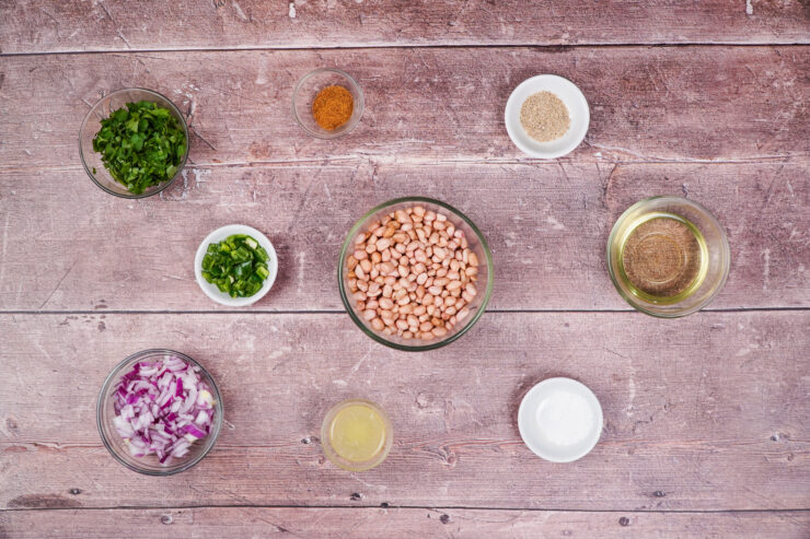 Overhead shot of the ingredients needed to make spicy cocktail peanuts including bowls of shelled peanuts, peanut oil, diced red onion, chopped cilantro, chopped jalapeno, lemon juice, salt, and pepper.