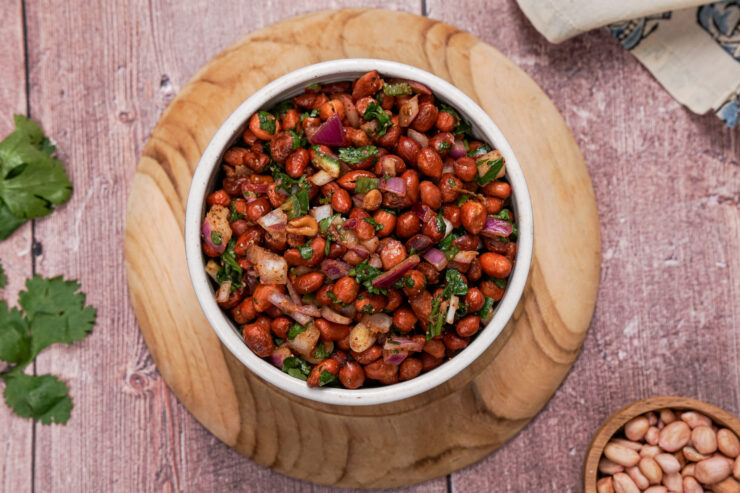 Overhead shot of a white bowl filled with spicy cocktail peanuts mixed with diced red onion and jalapenos, and chopped cilantro. It is sitting on a round wooden plate and a wooden bowl of plain peanuts is sitting in the bottom right corner.