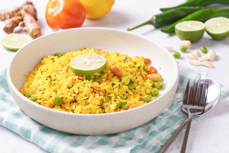 Curried poha rice in a white bowl and sitting on a blue and white plaid kitchen towel with a fork at its side