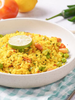 Curried poha rice in a white bowl and sitting on a blue and white plaid kitchen towel with a fork at its side