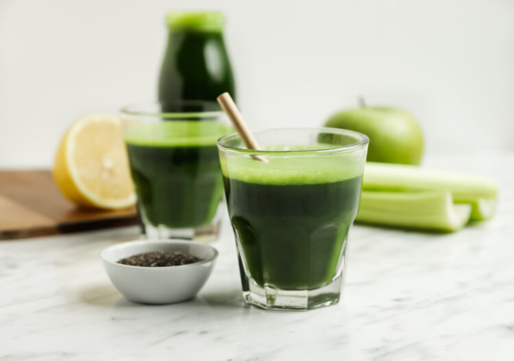 Two glasses of green juice with a lemon half, celery, apple, and bottle of green juice behind them