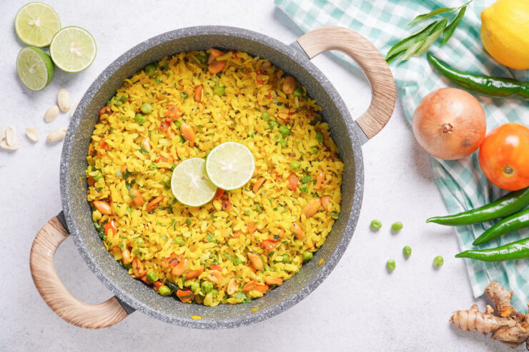 A pot of curried poha topped with slices of lime and surrounded by ingredients including onion, tomato, ginger and green peas.