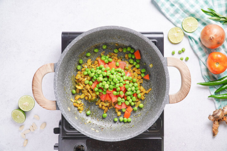 Ingredients for poha, including green peas, spices and tomatoes cook in a pot on an electric stovetop.
