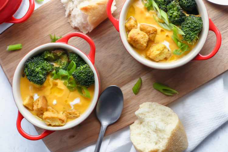 Two bowls of broccoli and carrot soup with bread on a wooden board.
