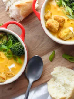 Two bowls of broccoli and carrot soup with bread on a wooden board.