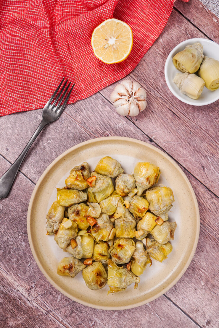 Baked artichoke hearts on a plate surrounded by ingredients including garlic, lemon and canned artichokes.
