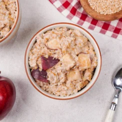 Two bowls of apple cinnamon oatmeal with an red apple sitting in front and a bowl of steel cut oats sitting behind on a red and white plaid kitchen towel.
