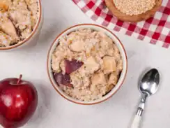 Two bowls of apple cinnamon oatmeal with an red apple sitting in front and a bowl of steel cut oats sitting behind on a red and white plaid kitchen towel.