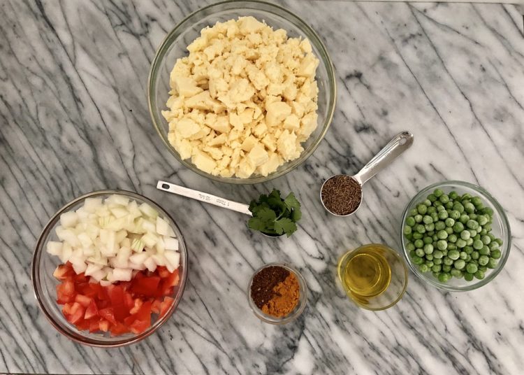 Bowl of crumbled paneer next to a bowl of chopped tomatoes and onion, measuring spoons full of spices, and a small bowl of chopped cilantro