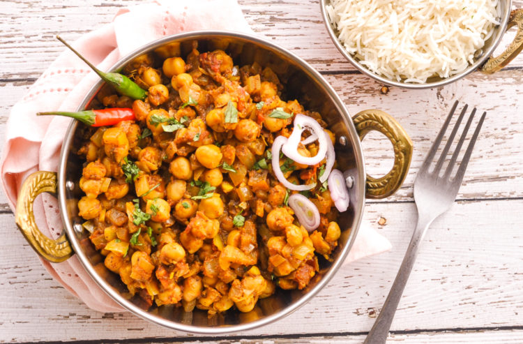 Pan of chickpea curry garnished with cilantro and sliced shallots with a fork and bowl of basmati rice on the side.