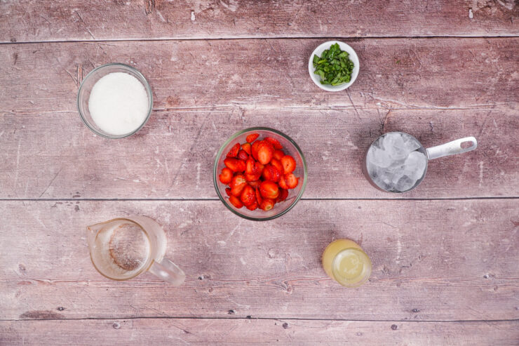 Overheat shot of bowls of ingredients to make the strawberry basil mocktail including sugar, basil, sliced strawberries, ice cubes, lemon juice, and seltzer.