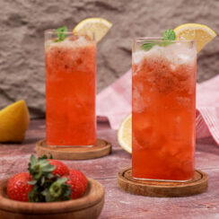 Two tall glasses of pink strawberry mocktail with lemon slices on the rim and sprigs of basil sitting in the drinks. They are sitting on wooden coasters with a wooden bowl of fresh strawberries sitting in front.