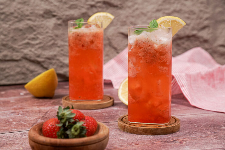 Two tall glasses of pink strawberry mocktail with lemon slices on the rim and sprigs of basil sitting in the drinks. They are sitting on wooden coasters with a wooden bowl of fresh strawberries sitting in front.