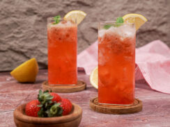 Two tall glasses of pink strawberry mocktail with lemon slices on the rim and sprigs of basil sitting in the drinks. They are sitting on wooden coasters with a wooden bowl of fresh strawberries sitting in front.