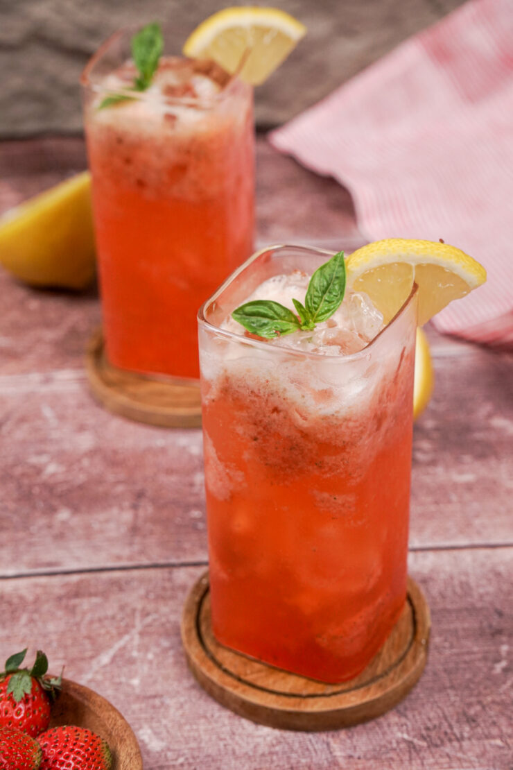 Tall glasses of fizzy pink strawberry basil mocktail with ice and a garnish of basil sprigs and lemon slices on the rims.