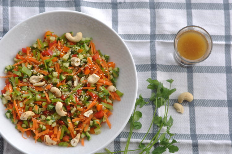 White bowl full of colorful Asian Vegetable Salad sitting on a white and black plaid kitchen towel with a small bowl of dressing on the side.