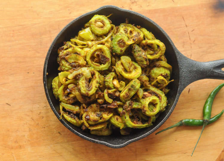 Small cast iron skillet full of sliced and spiced bitter gourd. It is sitting on a wooden surface with two long green chilies sitting next to it.