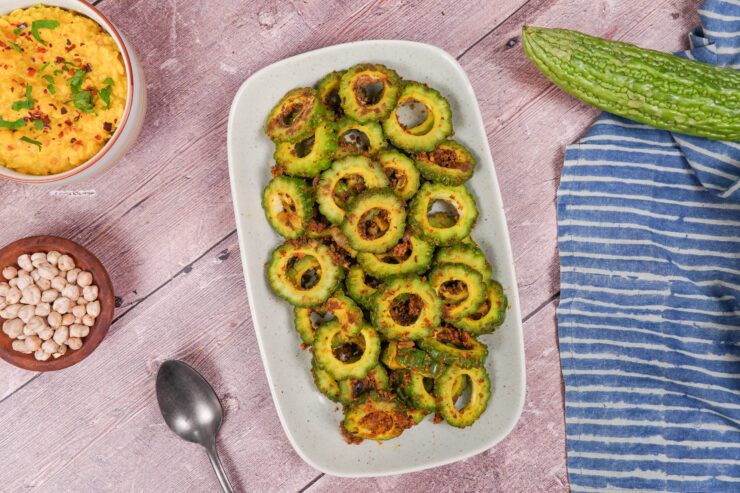Overhead view of a white rectangular platter with spiced bitter gourd slices on it. A whole bitter gourd is on its right side while there is a spoon and smaller bowl of fox nuts to the left.