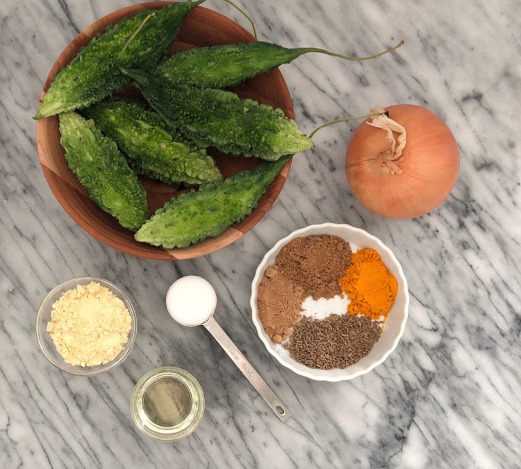 Overhead shot of a bowl of bitter gourds, an onion, a bowl of spices, a tablespoon of salt, a small bowl of oil, and a small bowl of garbanzo flour.