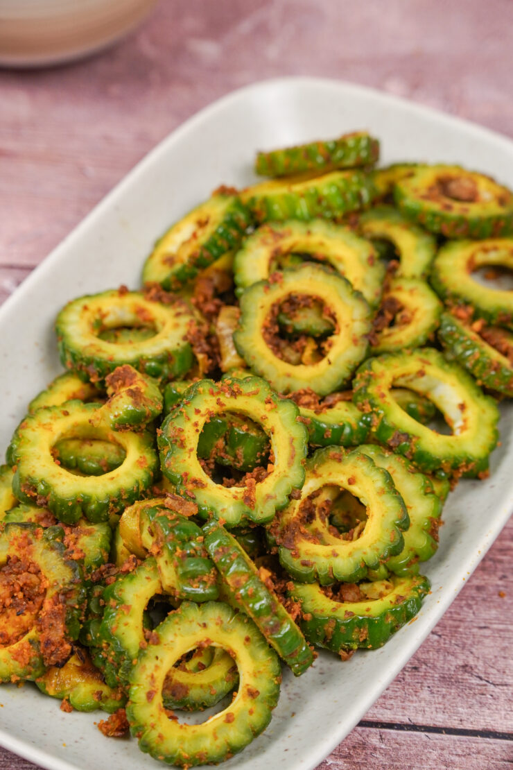 Closeup of sliced bitter gourd on a rectangular white plate. The bitter gourd is cooked and coated in garbanzo bean flour and spices.