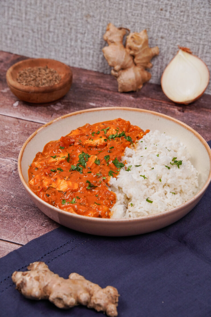 Bowl with red tofu tikka masala on one side and fluffy basmati rice on the other side, and chopped cilantro on top. There is a small wooden bowl of spices in the background along with a whole gingerroot and half of an onion. Meanwhile, a gingerroot sits in front as well.