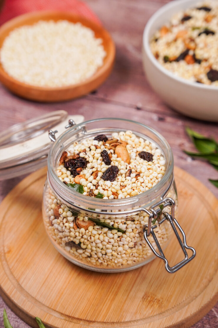 Jar of namkeen, which is a mixture of puffed millet, peanuts, raisins, sunflower seeds, and spices. It is sitting on a round wooden board with a white bowl of namkeen sitting behind.