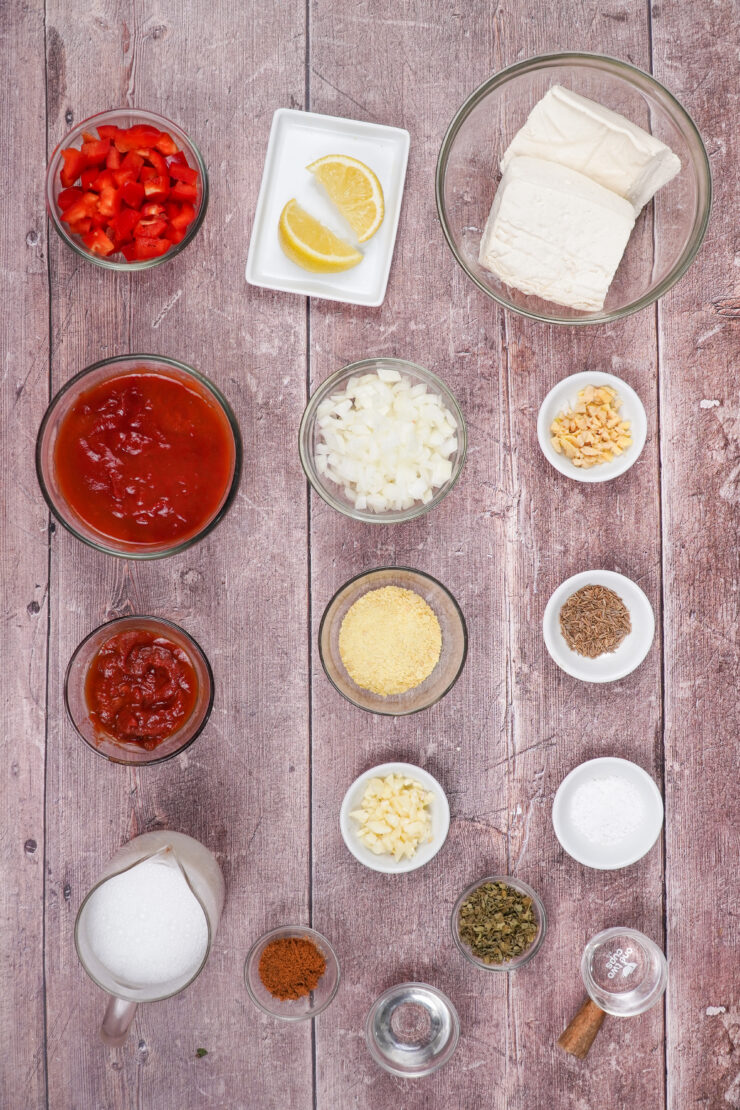 Overhead shot of bowls of the ingredients needed to make tofu tikka masala including diced red bell pepper, lemon wedges, blocks of tofu, tomato paste, diced onion, minced ginger, spices, chopped garlic, coconut milk, and white wine vinegar.