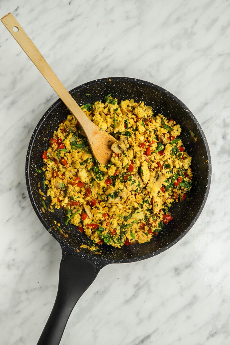 A wooden spoon mixes the ingredients in a pan full of curried tofu scramble with tomatoes, onion and tofu.